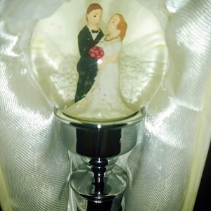 May include: A decorative bottle stopper featuring a clear glass orb with a miniature bride and groom figurine. The bride wears a white dress and the groom a black tuxedo. The stopper has a silver-colored metal base.