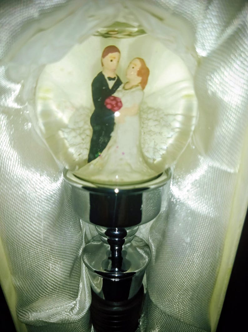May include: A decorative bottle stopper featuring a clear glass orb with a miniature bride and groom figurine. The bride wears a white dress and the groom a black tuxedo. The stopper has a silver-colored metal base.