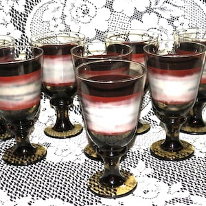 Puede incluir: Juego de ocho copas de vino de vidrio transparente con un diseño a rayas marrón, blanco y rojo. Las copas tienen una copa redonda y un tallo alto.