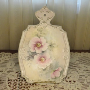 May include: A white ceramic cutting board with a floral design. The board features pink roses with green leaves and a white background. The board has a handle with a decorative design.