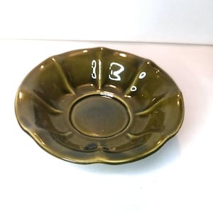 Könnte beinhalten: Eine grüne Keramikuntertasse mit gewelltem Rand. Die Untertasse hat eine zentrale, vertiefte Fläche und eine glänzende Oberfläche. Die Zahlen "313" spiegeln sich auf der Oberfläche. Das Gesamtdesign erinnert an eine Blume.