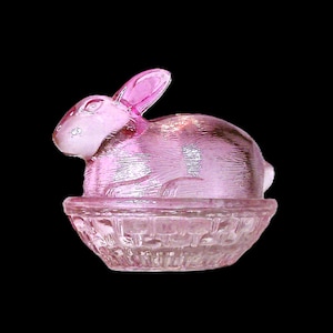 May include: A pink glass bunny figurine sitting in a basket-shaped dish. The bunny has long ears and is in a resting position. The dish and bunny are made of transparent glass, with a textured surface. The background is black.