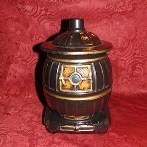 Puede incluir: Jarrón de galletas de cerámica negro y dorado con forma de estufa de leña. La estufa tiene una puerta con un diseño rojo, naranja y amarillo.