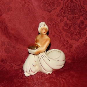 May include: A white ceramic figurine of a woman wearing a white head covering and a white dress with gold accents. She is holding a gold bowl in her hands. The figurine is sitting on a red background.