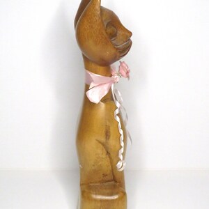 Puede incluir: Una figurita de gato de madera con una cinta rosa y una flor rosa alrededor del cuello. El gato est&aacute; sentado sobre una base de madera.