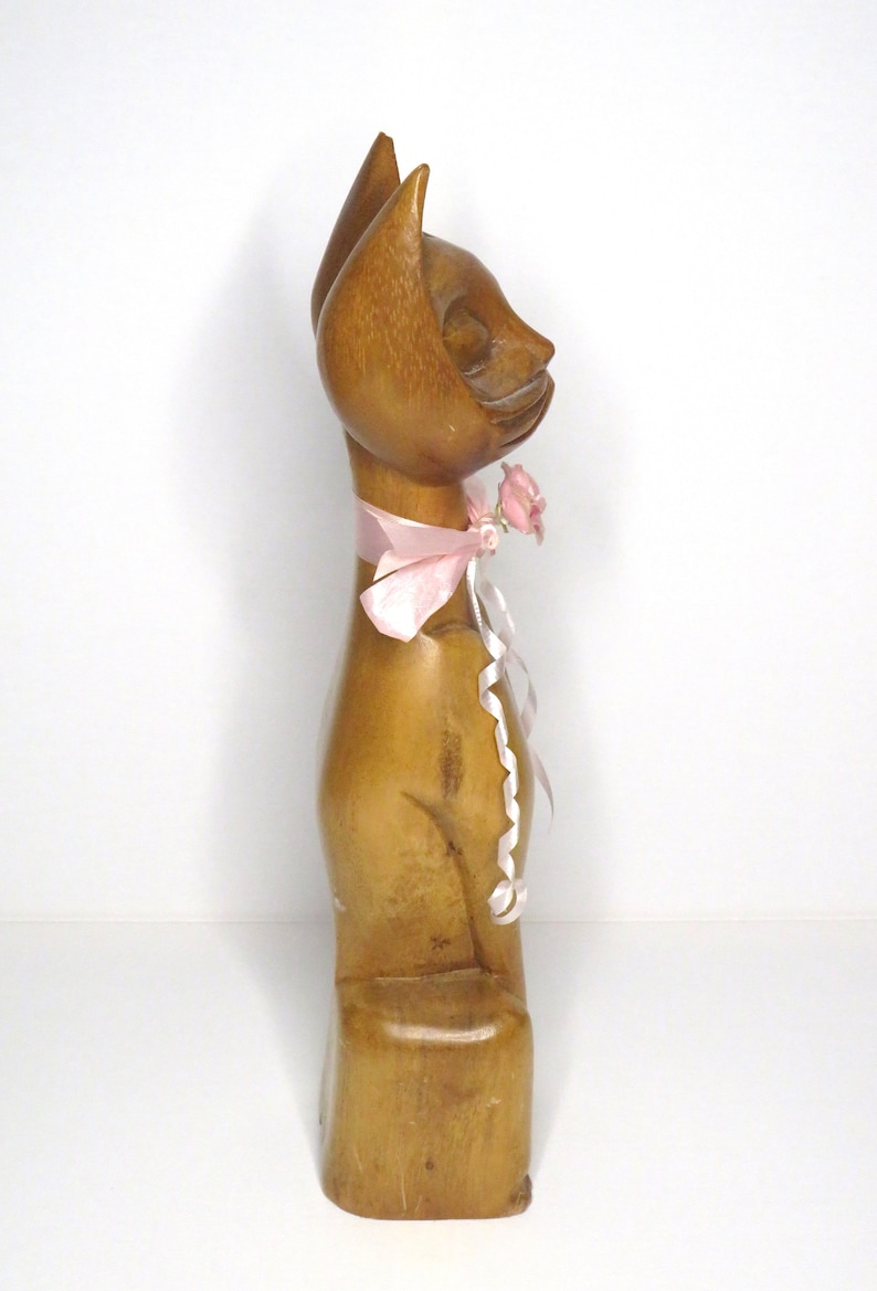 Puede incluir: Una figurita de gato de madera con una cinta rosa y una flor rosa alrededor del cuello. El gato est&aacute; sentado sobre una base de madera.