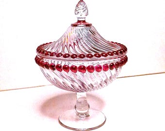 Puede incluir: Un bombonera de cristal transparente con tapa, con un diseño en espiral. El borde y el adorno de la tapa están adornados con cuentas rojas. El plato se apoya sobre una base transparente, ideal para presentar dulces.