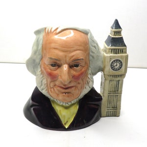 Könnte beinhalten: Eine Keramiktasse in Form eines Männerkopfes mit weißem Bart und schwarzer Jacke. Die Tasse steht neben einem weißen Keramikmodell von Big Ben.