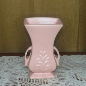Könnte beinhalten: Eine hellrosa Keramikvase mit zwei Henkeln und einem Blattdesign. Die Vase hat einen quadratischen Boden und eine breite Öffnung.