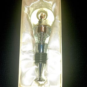 May include: A decorative bottle stopper with a chrome finish and a black rubber seal. The stopper features a clear, round top with a miniature wedding scene inside. Presented in a white box with satin lining.