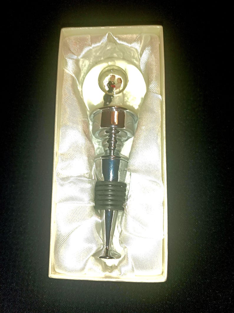 May include: A decorative bottle stopper with a chrome finish and a black rubber seal. The stopper features a clear, round top with a miniature wedding scene inside. Presented in a white box with satin lining.