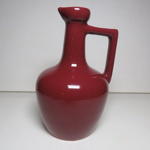 May include: A deep red ceramic pitcher with a wide base and a narrow neck. The pitcher has a handle on the side and a spout on the top.