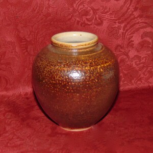 Könnte beinhalten: Eine kleine, braune Keramikvase mit einem gesprenkelten Überzug. Die Vase hat einen runden Körper und eine leicht ausgestellte Lippe.