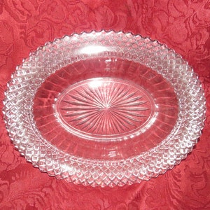 Puede incluir: Plato ovalado de vidrio transparente con un borde tallado en forma de diamante y un diseño de estrella en el centro. El plato está sobre una superficie roja estampada. Una pieza decorativa para servir comida.