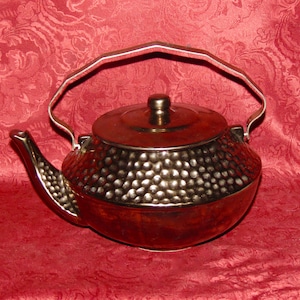 Puede incluir: Una tetera de color cobre con una textura martillada en el cuerpo y un asa plateada. La tetera tiene un pico curvo y una tapa con un pequeño pomo. El fondo es una tela roja estampada.