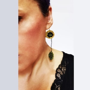 May include: A gold and black crocheted earring with a circular top and a dangling leaf-shaped bottom. The earring is attached to a black chain and is worn on the ear. The woman is wearing a black lace top.