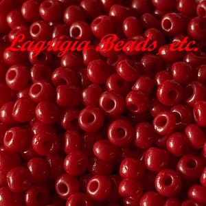 May include: A close-up image of a white bowl filled with small, round, red beads. The beads are all the same size and shape, and they are arranged in a random pattern. The bowl is sitting on a white surface.  Lagrigia Beads, etc.