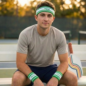 May include: A person wearing a white and green headband and wristbands with the text "Pickle Balls". The person is wearing a gray shirt and navy blue shorts. A pickleball paddle with colorful stripes rests nearby.