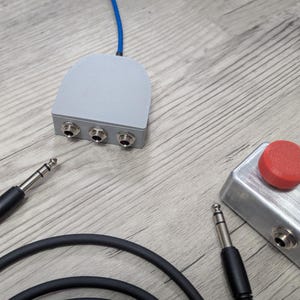 Puede incluir: Un divisor de audio gris con tres conectores de entrada y un cable azul. También se ve un pedal de metal plateado con un botón rojo, junto con dos cables de audio negros con conectores plateados. Los elementos están sobre una superficie de madera clara.
