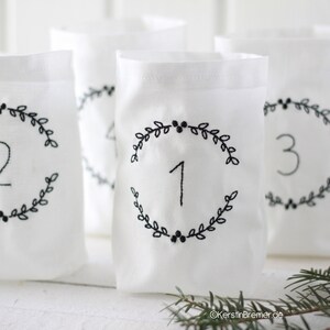 Könnte beinhalten: Drei weiße Stoffbeutel mit schwarzen gestickten Zahlen 1, 2 und 3 in einem Kranz aus Blättern. Die Beutel sind ein einfacher DIY-Adventskalender.
