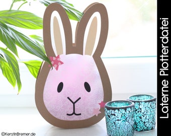 May include: A brown paper bunny lantern with pink accents and a white lace pattern. The lantern is lit up with a warm glow. Two teal blue glass candle holders are in the foreground.