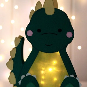 Pode incluir: Uma lanterna de papel verde em forma de dinossauro com espinhos amarelos e bochechas rosadas. A lanterna &eacute; iluminada por dentro com luzes brancas.