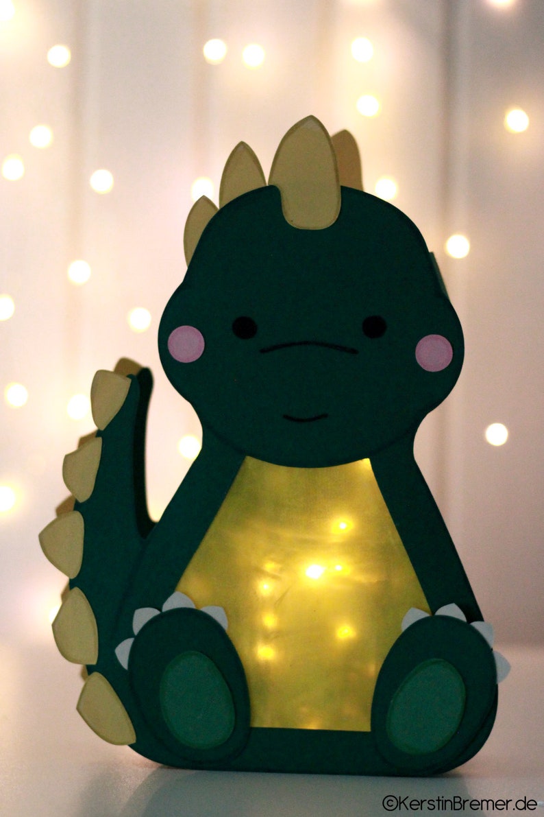 Pode incluir: Uma lanterna de papel verde em forma de dinossauro com espinhos amarelos e bochechas rosadas. A lanterna &eacute; iluminada por dentro com luzes brancas.