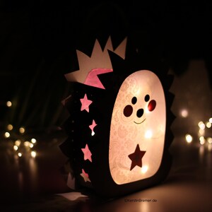 May include: A lantern shaped like a hedgehog with a black outer frame and a light-colored inner panel. The frame features cutouts of stars and spikes, while the inner panel displays a subtle pattern and a dark star. The lantern glows from within.