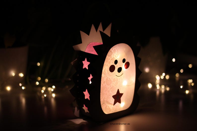 May include: A lantern shaped like a hedgehog with a black outer frame and a light-colored inner panel. The frame features cutouts of stars and spikes, while the inner panel displays a subtle pattern and a dark star. The lantern glows from within.