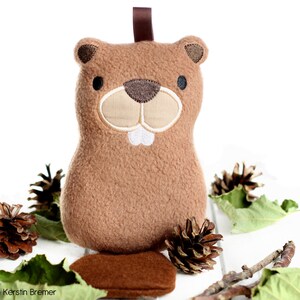 May include: A brown plush beaver toy with embroidered details, including a beige muzzle, white teeth, and black eyes. It has a brown ribbon loop at the top. The toy is surrounded by pine cones and leaves.