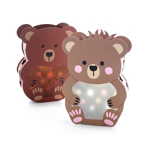 May include: Two bear-shaped paper lanterns with brown cardstock bodies. The front features a cutout belly area illuminated by internal multi-coloured lights. The bear on the right is light brown with pink cheeks, while the bear on the left is dark brown.