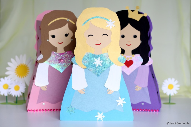 May include: Three paper dolls dressed as princesses. The princess on the left is wearing a pink dress with a sparkly bodice. The princess in the middle is wearing a light blue dress with a snowflake design. The princess on the right is wearing a purple dress with a red heart on the bodice.