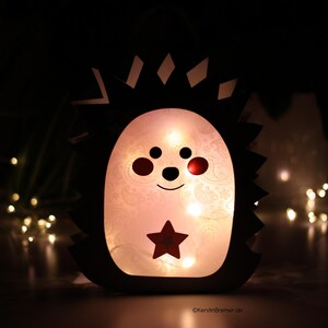 May include: A hedgehog-shaped lantern with a dark outer silhouette and a glowing, translucent white center. The face features two black eyes, a small nose, a curved mouth, and red cheeks. A red star is centered on the belly, illuminated from within.