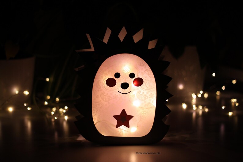May include: A hedgehog-shaped lantern with a dark outer silhouette and a glowing, translucent white center. The face features two black eyes, a small nose, a curved mouth, and red cheeks. A red star is centered on the belly, illuminated from within.