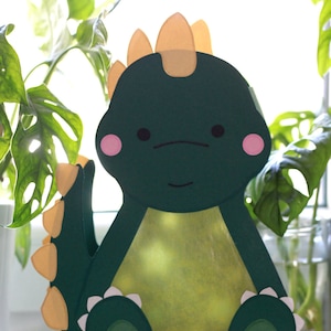 Pode incluir: Um dinossauro de papel verde com espinhos amarelos, bochechas cor-de-rosa e dentes brancos. O dinossauro est&aacute; sentado em uma superf&iacute;cie branca.