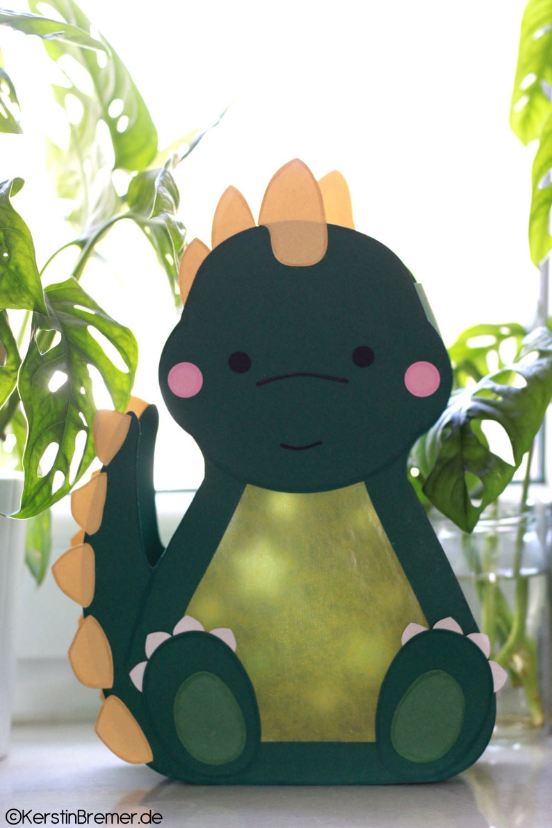 Pode incluir: Um dinossauro de papel verde com espinhos amarelos, bochechas cor-de-rosa e dentes brancos. O dinossauro est&aacute; sentado em uma superf&iacute;cie branca.