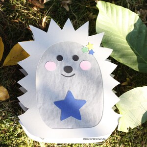 May include: A paper cutout of a hedgehog with a white spiky back and a light gray face. The face features two black circular eyes, a small black nose, a curved black mouth, and two pink circular cheeks. A blue star is centered on the belly.
