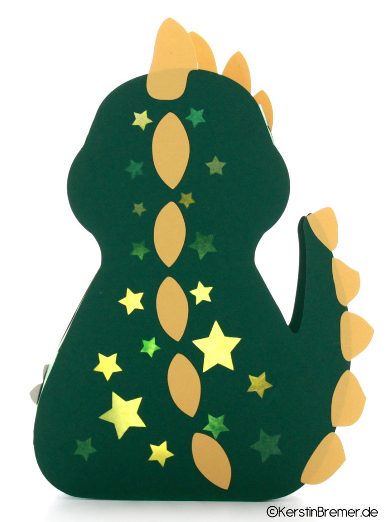 Pode incluir: Uma lanterna de papel verde em forma de dinossauro com uma espinha dorsal amarela e estrelas. A lanterna &eacute; decorada com estrelas amarelas e verdes.
