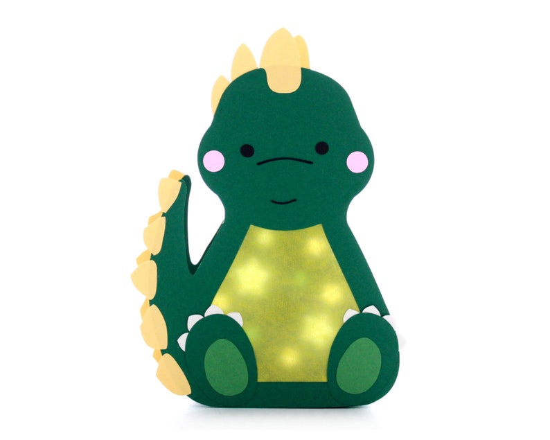 Pode incluir: Uma luz de noite de dinossauro verde com espinhos amarelos e bochechas rosadas. O dinossauro est&aacute; sentado com as pernas cruzadas e tem uma luz amarela que brilha atrav&eacute;s do seu corpo.