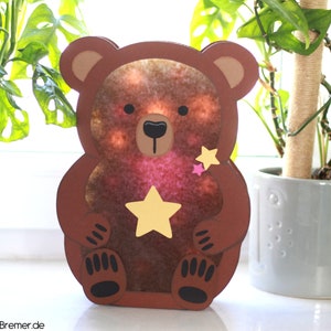 Könnte beinhalten: Ein brauner Papierbär mit einem gelben Stern in seinen Pfoten und einem rosa Stern an der Seite. Der Bär ist eine Leuchtlaterne mit einem durchscheinenden Papierkörper.