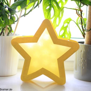 May include: A yellow star-shaped paper lantern with a white interior. The lantern is sitting on a windowsill in front of two potted plants.