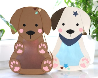 May include: Two dog-shaped paper lanterns. One is brown with star accents, the other is white with a brown ear and blue accents. Both have pink cheeks and paws, and black noses and eyes. The lanterns are sitting on a white surface.