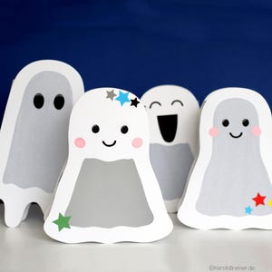 May include: Four white paper ghosts with different facial expressions and colorful star decorations. The ghosts are standing on a white surface against a blue background.