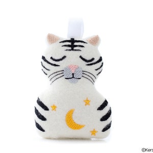Könnte beinhalten: Weißes Plüsch-Tiger-Spielzeug mit schwarzen Streifen und gestickten Details, darunter geschlossene Augen, eine rosa Nase und einen gelben Halbmond mit Sternen.
