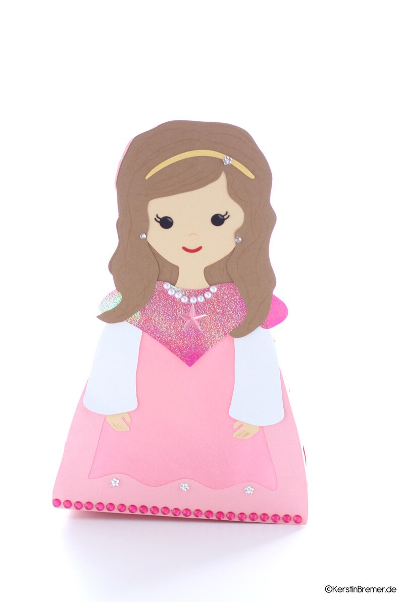 May include: A pink princess-themed party favor box with a cutout of a girl with brown hair, wearing a pink dress, a gold headband, and a pearl necklace. The box has a sparkly pink and white design and a row of pink rhinestones around the bottom.