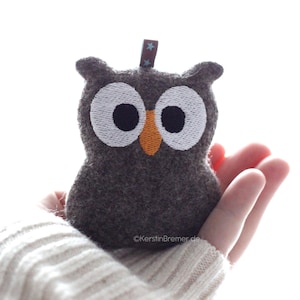 May include: A small, dark grey felt owl plush toy featuring large white embroidered eyes with black pupils and an orange stitched beak. A brown fabric tab with small blue star patterns is attached to the top of the owl's head. The text KerstinBremer.de is visible.