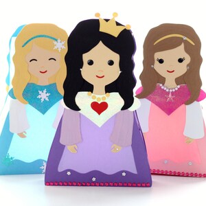 May include: Three paper dolls dressed as princesses. The princess on the left is wearing a light blue dress with a snowflake design. The princess in the middle is wearing a purple dress with a red heart and a gold crown. The princess on the right is wearing a pink dress with a gold star and a gold headband.