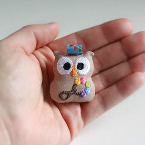 以下が含まれることがあります： 刺繍された目で虹色のガラガラを持つ、小さな茶色のフクロウのぬいぐるみ。フクロウは手のひらに乗っています。
