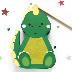 Pode incluir: Um dinossauro de papel verde com uma crista amarela, espinhos amarelos na cauda e uma barriga amarela. O dinossauro tem um rosto amig&aacute;vel com olhos pretos e bochechas rosadas. O dinossauro est&aacute; sentado em um fundo branco com estrelas espalhadas.