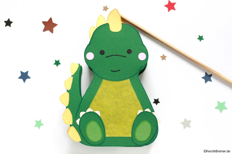Pode incluir: Um dinossauro de papel verde com uma crista amarela, espinhos amarelos na cauda e uma barriga amarela. O dinossauro tem um rosto amig&aacute;vel com olhos pretos e bochechas rosadas. O dinossauro est&aacute; sentado em um fundo branco com estrelas espalhadas.